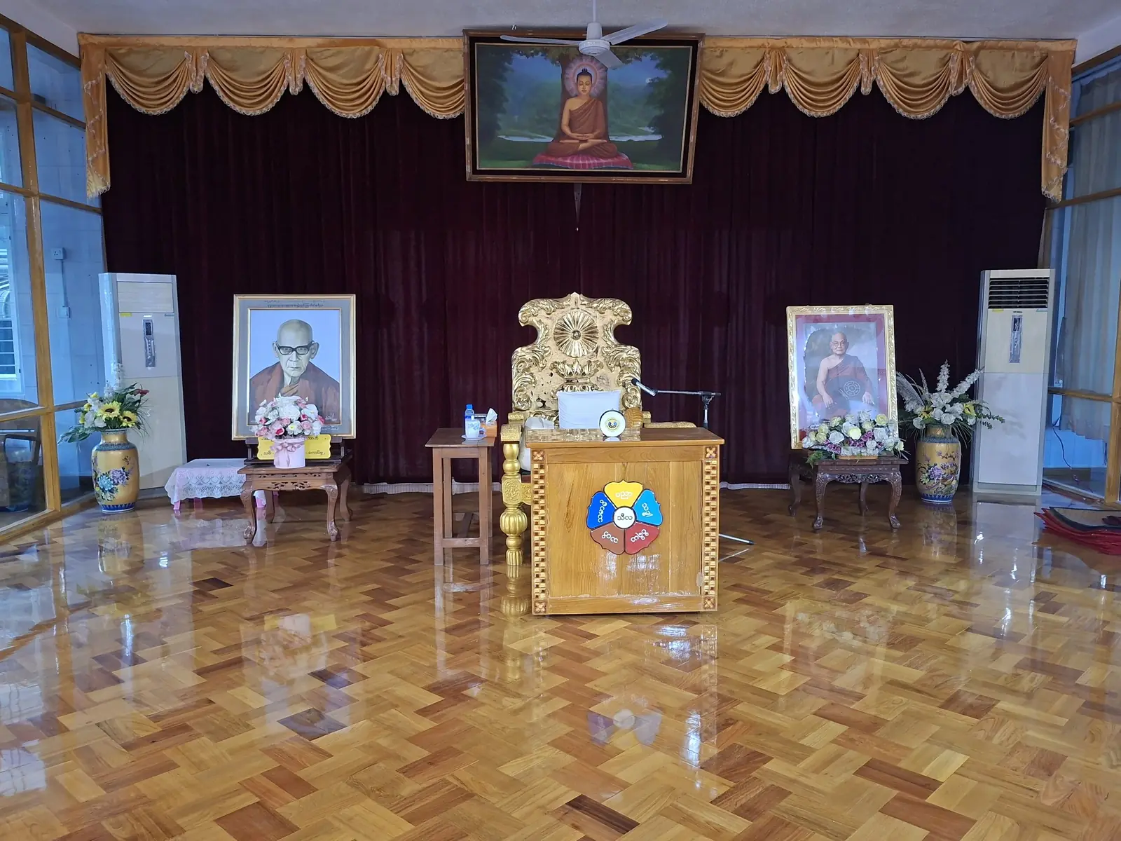 An empty Dhamma hall with a gleaming parquet floor, a gilded teacher's seat and lectern, framed portraits of two senior monks on either side and a painting of the seated Buddha above