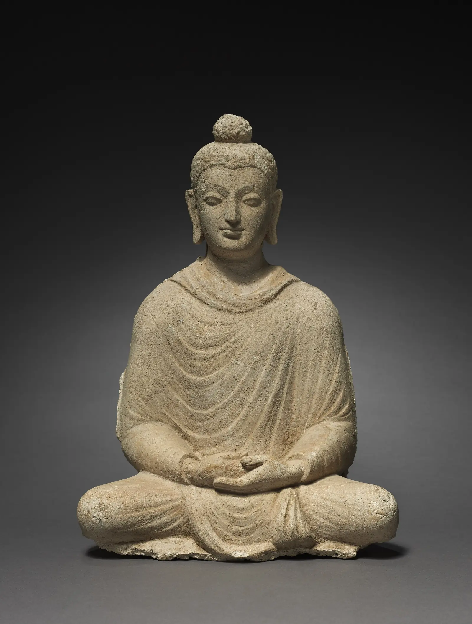 Stone sculpture of the Buddha seated cross-legged in meditation, hands resting in his lap, from Hadda in ancient Gandhara, photographed against a dark grey background